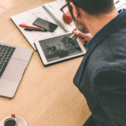 Mann sitzt am Schreibtisch und arbeitet auf Tablet; neben ihm Büromaterialien und ein Laptop