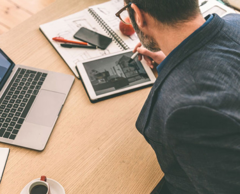 Mann sitzt am Schreibtisch und arbeitet auf Tablet; neben ihm Büromaterialien und ein Laptop