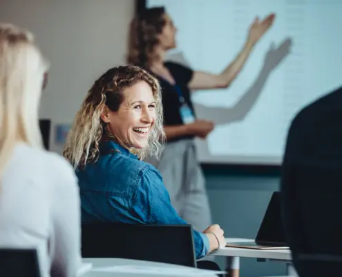 Rückansicht einer Frau, die im Publikum sitzt, während einer Konferenz zu einem Kollegen blickt und lächelt.