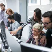 2 Frauen und 1 Mann schauen auf einen Computerbildschirm, im Hintergrund 2 Männer und 1 Frau