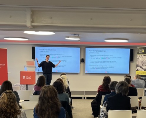 Vortragende, im Publikum Frauen, im Hintergrund elektronische Tafeln