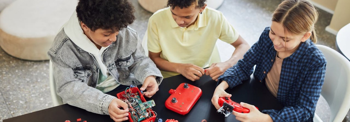 3 Kinder sitzen an einem schwarzen Tisch und arbeiten an kleinen roten Roboter-Modellen.