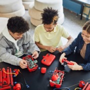 3 Kinder sitzen an einem schwarzen Tisch und arbeiten an kleinen roten Roboter-Modellen.