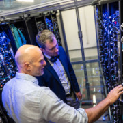 2 Männer stehen in einem technisch ausgestatteten Raum zwischen dicht verkabelten Geräten und Schaltschränken.